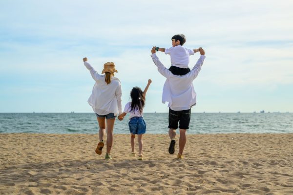 Family of four at the beach, seen from behind, with adults raising arms as a child sits on a dad's shoulders near the ocean.