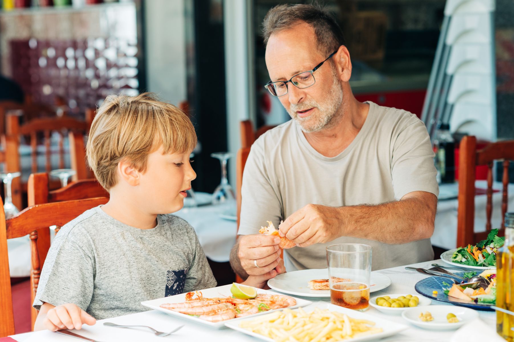 Man and boy dining at a restaurant, peeling shrimp at a table with plates of seafood and sides nearby.