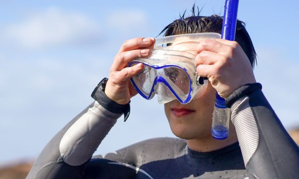 Person adjusting a blue snorkeling mask and snorkel against a clear sky, wearing a wetsuit.
